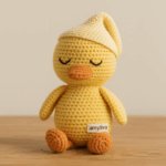 "Handmade amigurumi duck by AMYLIVA, featuring soft yellow cotton yarn, an orange beak and feet, sleepy embroidered eyes, and a cream nightcap, sitting on a light wooden surface."