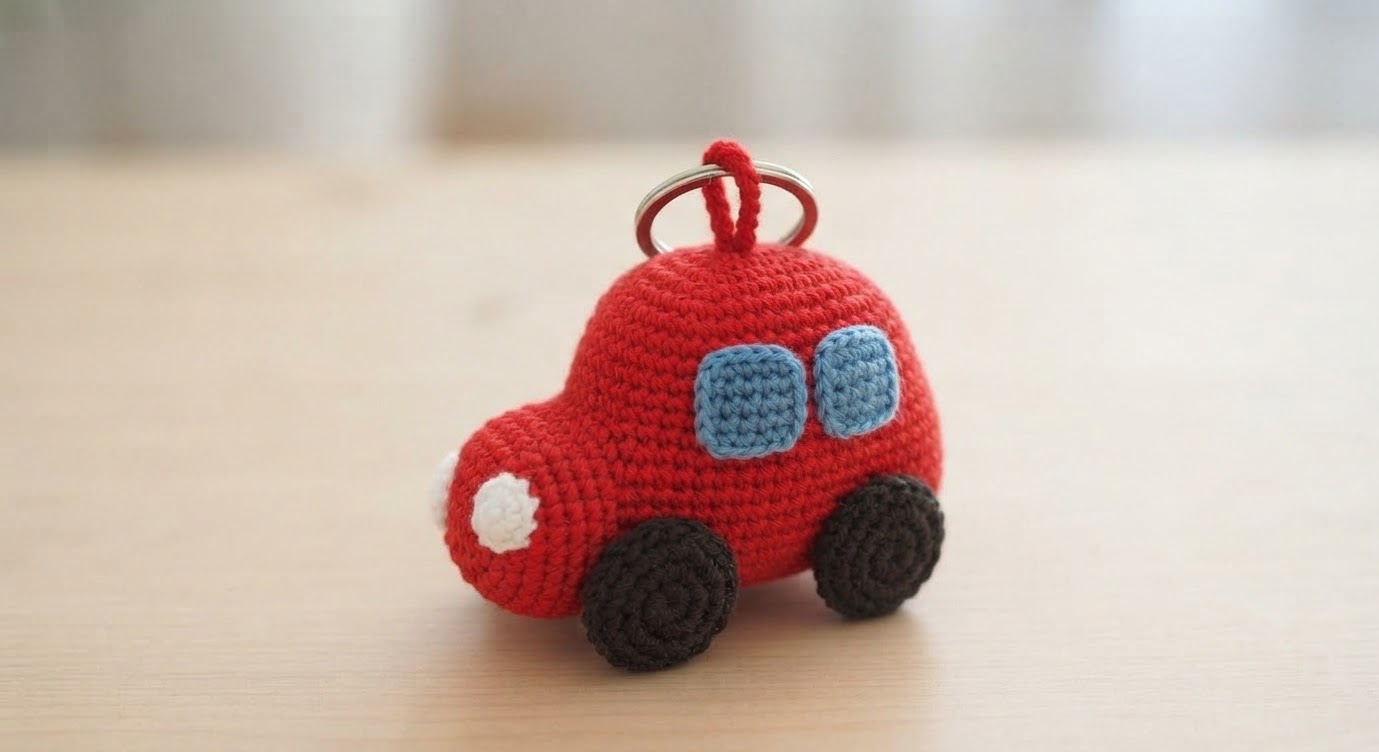 Mini crochet car keychain with a red body, blue windows, white headlights, and small dark wheels.