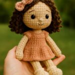 Handmade amigurumi doll with curly brown hair, blush bow, and peach dress sitting in a human hand outdoors, surrounded by green garden foliage.