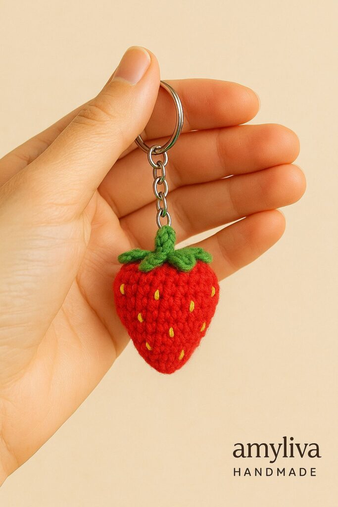 Handmade crochet strawberry keychain with red yarn body, yellow seed stitches, and green leafy top. Held in hand, with “amyliva HANDMADE” text in the corner.
