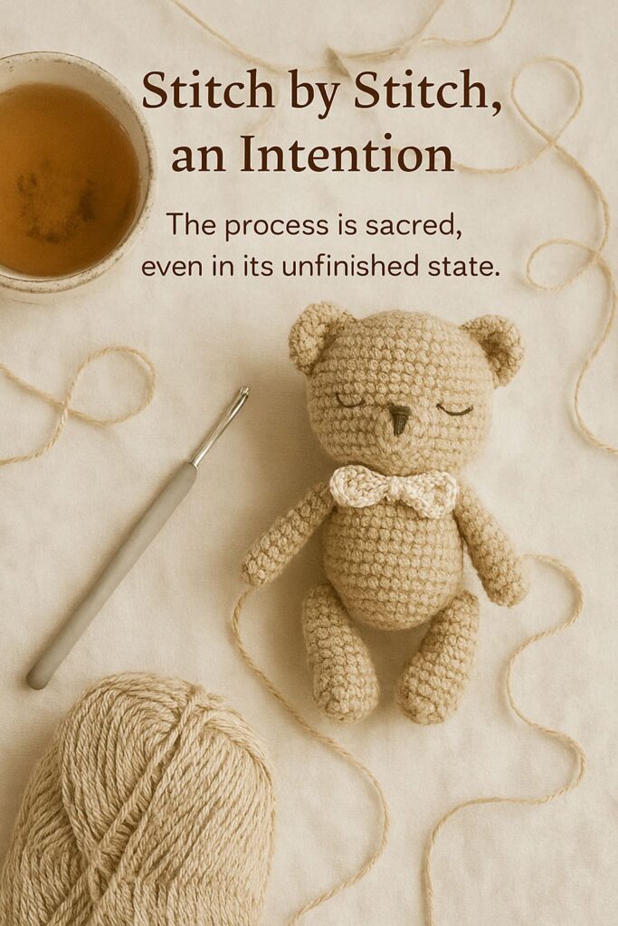 A crocheted teddy bear with a bow tie lies on a soft cream fabric, surrounded by a ball of yarn, a crochet hook, and a cup of tea. A loose strand of yarn gently frames the scene. The warm, neutral tones create a peaceful, meditative atmosphere. Text reads: “Stitch by Stitch, an Intention. The process is sacred, even in its unfinished state.